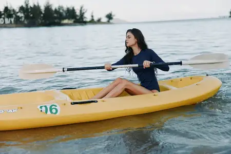 Молодая женщина гребёт на жёлтом каяке по спокойной воде на фоне облачного неба и естественного ландшафта, одетая в тёмно-синий костюм для занятий водными видами спорта и приключений на открытом…