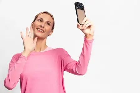 Счастливая молодая женщина в розовом свитере держит смартфон и машет рукой, делая селфи