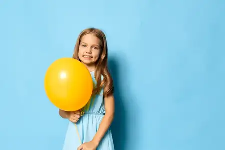 Маленькая девочка в голубом платье держит жёлтый воздушный шар на фоне ярко-голубого фона