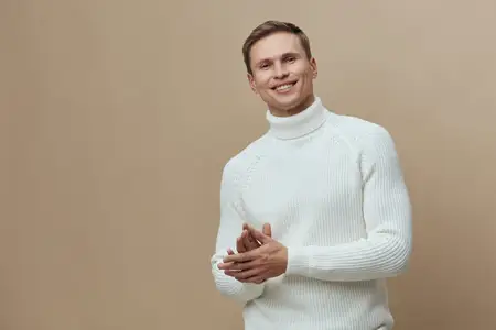Улыбающийся молодой мужчина европеоидной расы в белом вязаном свитере смотрит в камеру со сложенными руками, позируя изолированно на бежевом фоне