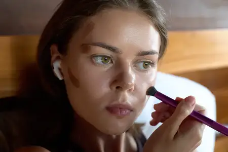Крупный план: женщина наносит макияж кисточкой, видны патчи на лице, беспроводной наушник