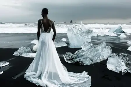 Невеста в белом свадебном платье стоит на черном песчаном пляже, на заднем плане айсберги на фоне захватывающего дух пейзажа