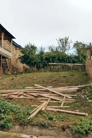 Заброшенный двор с разбросанными деревянными досками возле старого здания, заросшая трава и сломанный забор