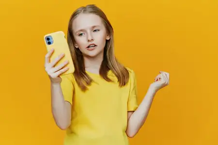 Веселая маленькая девочка в ярко-желтой рубашке с таким же смартфоном, демонстрирующая уверенность и игривость на фоне живого желтого фона