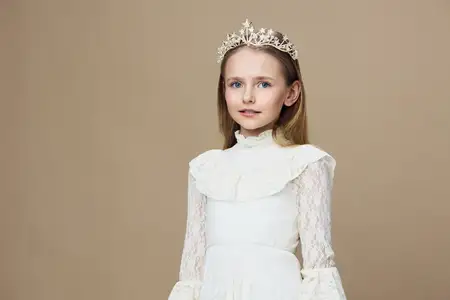 Портрет радостной маленькой девочки в белом кружевном платье и короне, стоящей на фоне мягкого бежевого фона