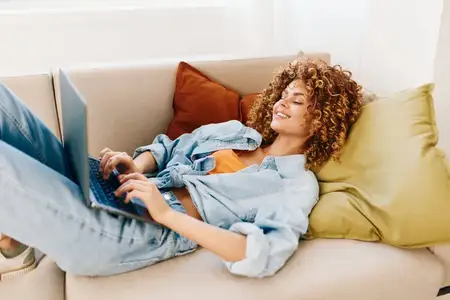 Улыбающаяся женщина работает за ноутбуком в уютном домашнем офисе, окружённая современными технологиями и комфортной обстановкой гостиной