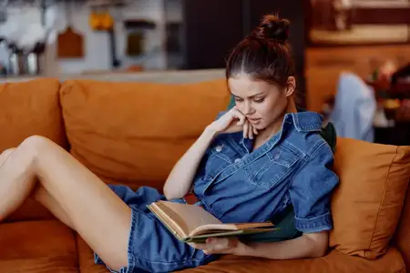 Женщина в джинсовом платье сидит на диване, скрестив ноги, и читает книгу в комфортной домашней обстановке