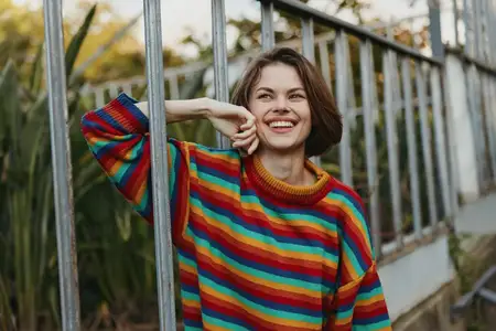 Улыбающаяся женщина в полосатом свитере и повседневной одежде прислонилась к перилам теплицы в осеннем саду на улице