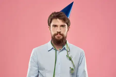 Бородатый мужчина с тортом на розовом фоне, на голове у него праздничный колпак
