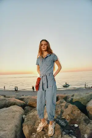 Женщина в стиле lifestyle, одетая в современный бохо-вестерн деним стритстайл, уверенно позирует на каменистом берегу на закате, демонстрируя джинсовый комбинезон casual и кроссовки в тёплой…