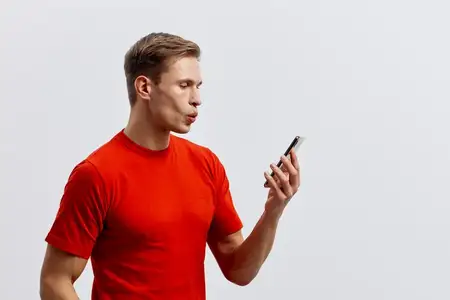 Мужчина в повседневной красной футболке держит смартфон, сосредоточенно глядя на экран, изолированный на нейтральном белом фоне
