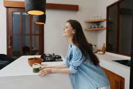 Молодая женщина улыбается, опираясь на кухонную стойку, и наслаждается зеленым смузи