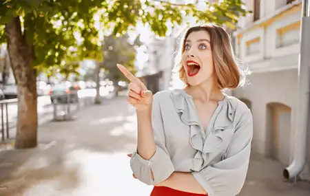 Счастливая блондинка с ярким макияжем жестикулирует руками возле здания, модная одежда, стиль