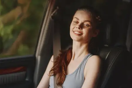Улыбающаяся молодая женщина в повседневной одежде сидит в машине на фоне яркой природы, излучая расслабленную летнюю атмосферу, идеально подходящую для тем образа жизни и путешествий
