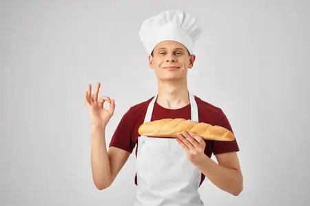 Повар-мужчина в белом фартуке держит в руках буханку хлеба