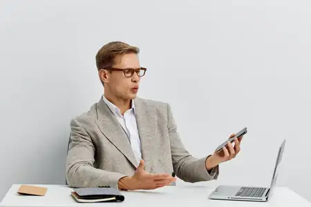 Удивленный мужчина использует смартфон и ноутбук для удаленной работы цифрового кочевника, в очках и повседневной куртке на однотонном белом фоне