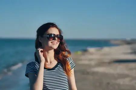 Портрет красивой путешественницы в очках, с рыжими волосами и в футболке на фоне пляжного летнего пейзажа с морем