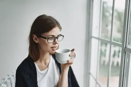 Чашка кофе в руках женщины, сидящей в кресле у окна в интерьере