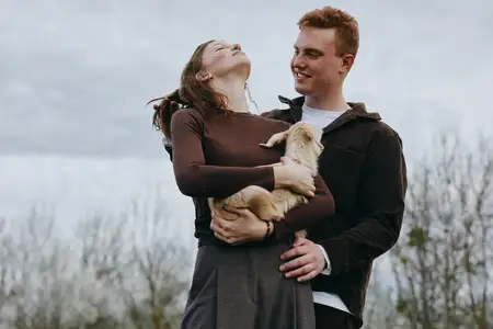 В естественном природном окружении молодая женщина с радостью обнимает козленка, в то время как молодой человек смотрит на них с улыбкой, оба одеты в повседневную осеннюю одежду, позади них голые…
