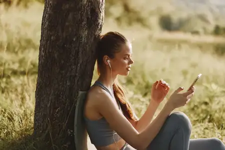 Молодая женщина в спортивной одежде слушает музыку через наушники, сидя у дерева на пышной зелёной лужайке