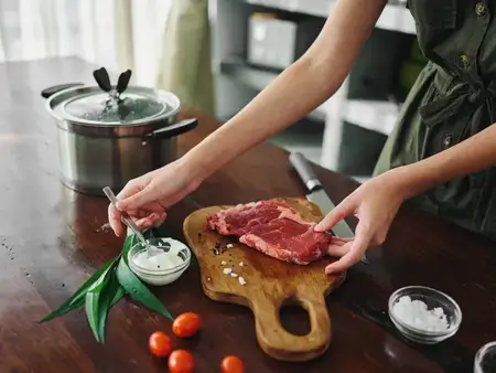 Женщина на кухне готовит ужин из мяса и овощей со специями на деревянном столе на фоне стильной кухни