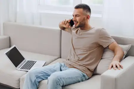 Улыбающийся мужчина ведет телефонный разговор, сидя на уютном диване с ноутбуком рядом, что отражает современную атмосферу домашнего офиса