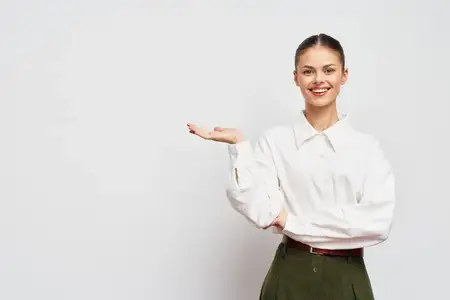 Жизнерадостная женщина в белой рубашке и зеленых брюках улыбается, протягивая руку, создавая пространство для текста или сообщений