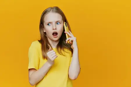 Маленькая девочка с удивленным выражением лица разговаривает по желтому телефону, одетая в повседневную желтую рубашку, на фоне ярко-желтого фона, что идеально подходит для ярких и игривых концепций
