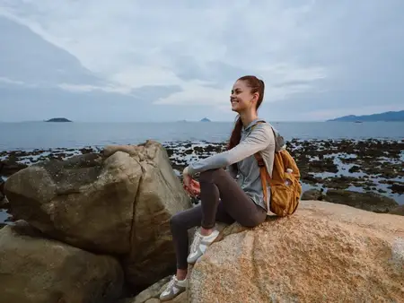Жизнерадостная молодая женщина в спортивной одежде сидит на камне у моря, излучая радость и безмятежность на фоне живописного прибрежного пейзажа с мягкими цветами