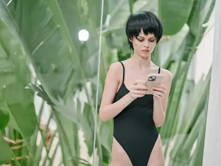 Женщина в чёрном купальнике, увлечённая своим смартфоном, стоит среди крупных зелёных тропических листьев, передавая ощущение расслабленности и цифровой вовлечённости