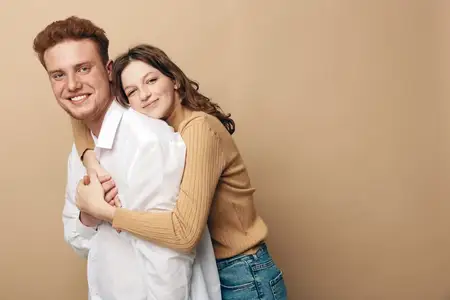 Счастливая молодая пара в повседневной одежде тепло обнимается на фоне однотонного бежевого фона