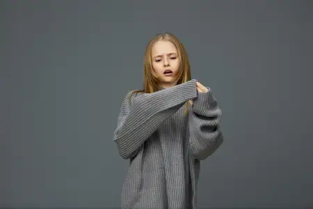 Юная модель со светло-каштановыми волосами в свободном сером объемном свитере вызывает задумчивые эмоции на фоне приглушенного серого фона