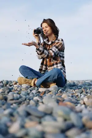 Сцена уличной фотосъемки: женщина-фотограф на галечном пляже сосредоточена на кадрировании снимка с камерой в руках, среди брызг и ветра