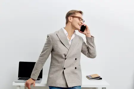 Улыбающийся мужчина разговаривает по смартфону в повседневном пиджаке, стоя рядом с ноутбуком на белом фоне, демонстрируя счастье и вовлеченность в цифровой кочевой образ жизни и удаленное общение