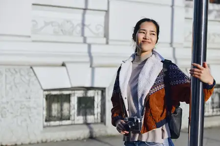 Непринуждённый снимок: улыбающаяся женщина с фотоаппаратом рядом со столбом на залитой солнцем улице