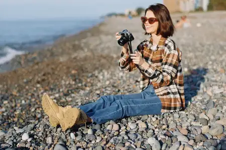 Портретный кадр фотографа в повседневной одежде на каменистом пляже, с камерой в руках, в клетчатой рубашке и джинсах, наслаждающегося солнечным светом, волнами и настроением