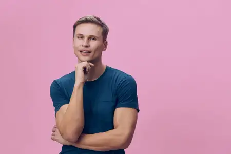 Счастливый молодой человек с blond-волосами, одетый повседневно в синюю футболку, стоит на фоне розовой стены, выглядит уверенным и жизнерадостным