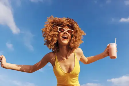 Жизнерадостная молодая женщина в солнцезащитных очках и жёлтом купальнике держит освежающий напиток, излучая счастье в яркой обстановке на открытом воздухе