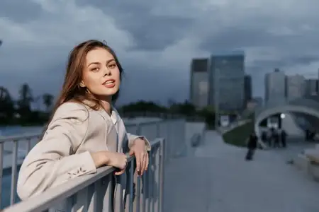 Молодая женщина в бежевом пальто, опирающаяся на перила снаружи на фоне городского пейзажа под облачным небом, демонстрирующая городской модный образ жизни, повседневный стиль, уверенное настроение