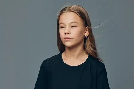 Маленькая девочка с прямыми волосами, одетая в однотонную черную майку, смотрит в сторону от камеры с расслабленным лицом и сомкнутыми губами