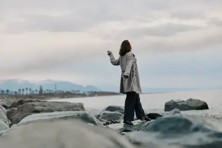 Человек в пальто стоит на каменистом берегу у моря под облачным небом