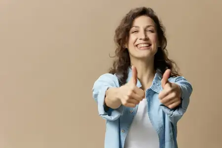 Счастливая улыбающаяся кудрявая красивая женщина в джинсах и повседневной рубашке показывает жест «большой палец вверх» (Like) в камеру, позируя изолированно на бежевом пастельном фоне