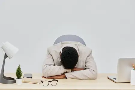 Молодой человек испытывает стресс или разочарование на работе, сидя за столом с опущенной головой и руками на голове