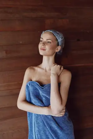 Молодая женщина, завернутая в синее полотенце и в шапочке для душа, спокойно стоит у деревянной стены, излучая безмятежность и заботу о себе в уютной домашней обстановке
