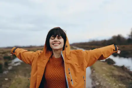 Женщина на открытом воздухе в стиле лайфстайл в оранжевой куртке с капюшоном: улыбается, раскинув руки у воды, осенний пейзаж с облачным небом и рекой, естественный непринужденный портрет в теплом…