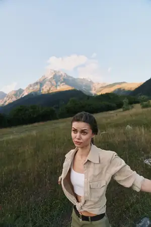 Улыбающаяся, уверенная женщина наслаждается отдыхом на природе, горы на заднем плане, в бежевой рубашке и куртке, открытая поза, свежий воздух, природа, свобода и исследования в сиянии яркого пейзажа
