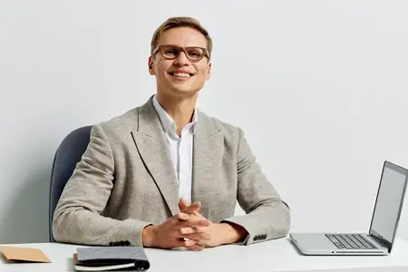 Улыбающийся мужчина европеоидной расы работает с ноутбуком в современном светлом офисе, в очках, легком костюме и белой рубашке, смотрит в камеру с уверенным счастливым выражением, сидя за столом
