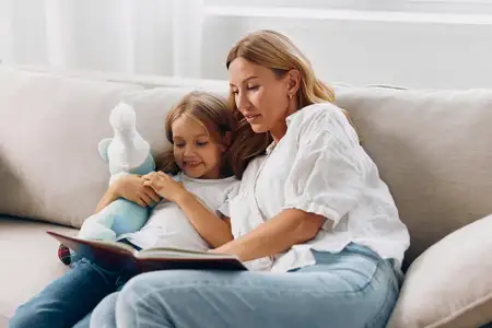 Мать и дочь общаются на диване, читая книгу вместе и держа плюшевого мишку