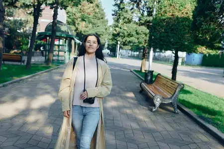 Непринужденная прогулка на свежем воздухе по солнечному парку, с камерой в руке, развевающимся пальто и безмятежным выражением лица, когда природа окружает спокойный пешеходный момент
