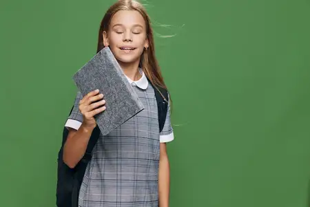 Ученица в школьной форме и с рюкзаком позирует с серым чехлом для планшета на зеленом фоне, что символизирует обучение и использование цифровых устройств